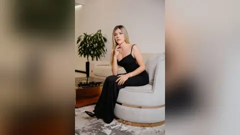 Elegant woman in a black evening gown sitting on a grey armchair in a modern minimalist living room.