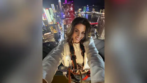 Smiling woman in white jacket and safety harness overlooking a glowing city skyline at night.