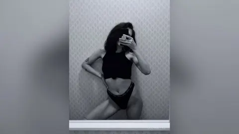 Moody black and white mirror selfie of a tattooed woman in a black crop top and briefs against a patterned wall.