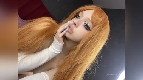 Young woman with long strawberry blonde hair and dramatic lashes wearing a white top in a soft, moody setting.