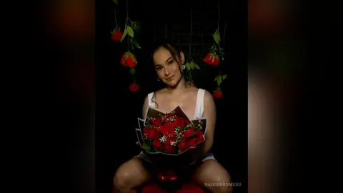 NahomiRomero poses in a white tank top, holding a large bouquet of red roses against a dark background.