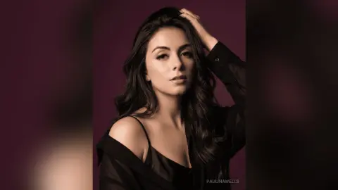 Elegant woman with long wavy hair wearing a black silk outfit against a moody dark purple background.