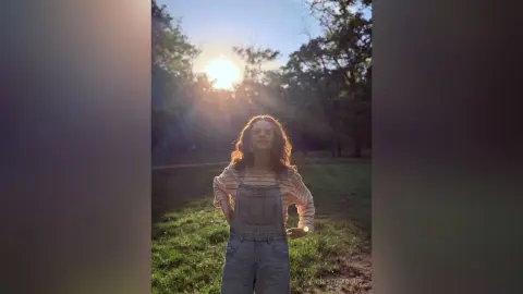 Young woman in denim overalls standing in a sunlit forest during a dreamy golden hour sunset.