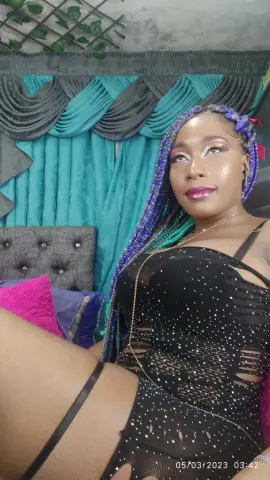 Smiling Black woman with long blue braids wearing a black sequined cutout dress in a bedroom setting.