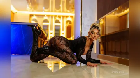 Anastasia poses on a light floor wearing a black lace bodysuit and black high heels.