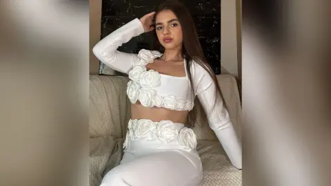 Young woman in a white floral crop top and matching skirt sitting on a couch in a stylish indoor setting.