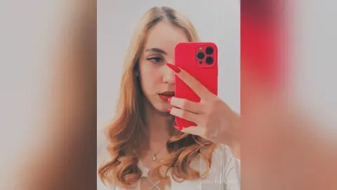 Blonde woman in a white top taking a red phone selfie with long red nails in a bright indoor setting.