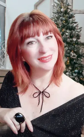 Smiling woman with red hair in a black sparkly dress posing near a festive Christmas tree.