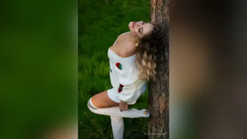 Blonde woman in a white strawberry sweater leaning against a tree in a lush green meadow.