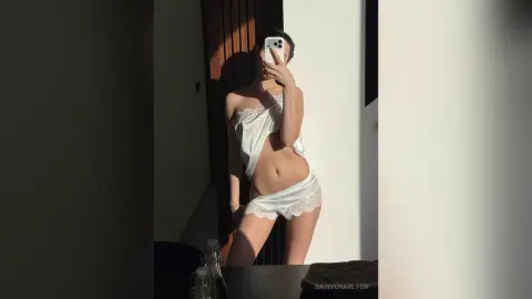 Young woman in white lace lingerie set posing for a mirror selfie in warm sunlight.