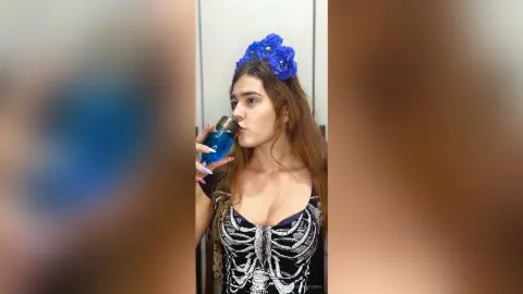Young woman with blue flower crown drinking from a blue water bottle in a casual indoor setting.