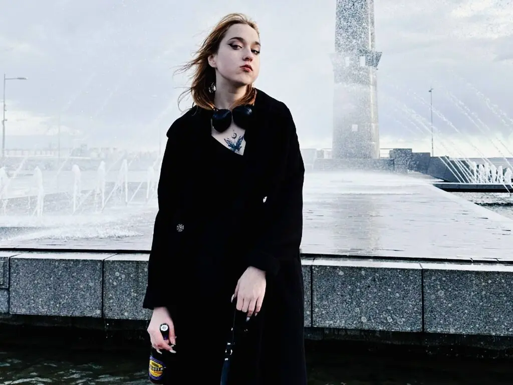 OksiKoxy stands outdoors in a black coat with her hair blowing in the wind near a large water fountain.