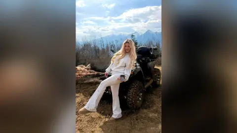Anita stands outdoors in a white long-sleeve top and white flared pants against a mountain landscape.