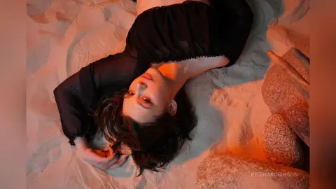 Young woman in a black dress relaxing on white sand under warm sunset lighting.