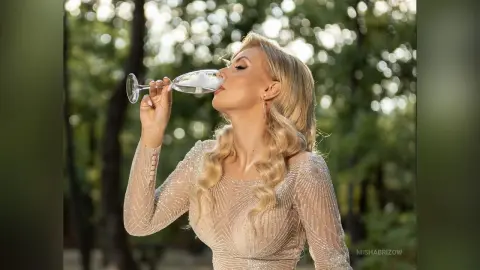 Elegant blonde woman in a sparkling sequin gown sipping champagne in a sunlit forest setting.