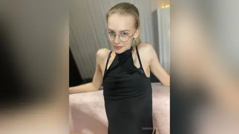 Elegant woman in a black halter neck dress and glasses posing in a bright, minimalist indoor setting.
