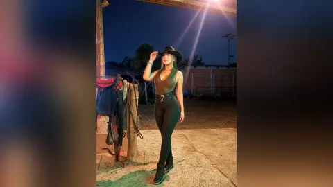 Stylish woman in a black sleeveless top and leggings wearing a cowboy hat in a rustic outdoor setting at night.