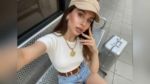 Stylish woman in white t-shirt and denim shorts sitting on a bench with silver luggage, ready for travel.