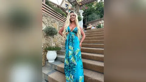 Anita stands on stone stairs wearing a long, blue tropical print sundress with a deep plunging neckline.