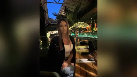 Young woman in a black blazer sitting at a restaurant table with tropical plants and warm evening lighting.