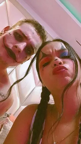 Darly_AndDixon and her partner pose close together, showing off their heavy, dark eye makeup and facial tattoos against a white padded wall.