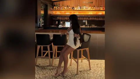 Elegant woman in a white mini dress sitting at a sophisticated bar with warm lighting.