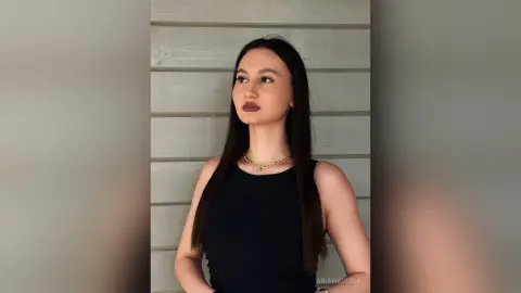 Elegant woman in a black sleeveless top and gold necklace posing against a neutral wood paneled wall.
