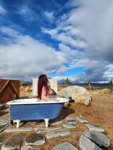 Redheaded woman relaxing in a vintage blue clawfoot bathtub outdoors under a cloudy sky in a desert landscape.