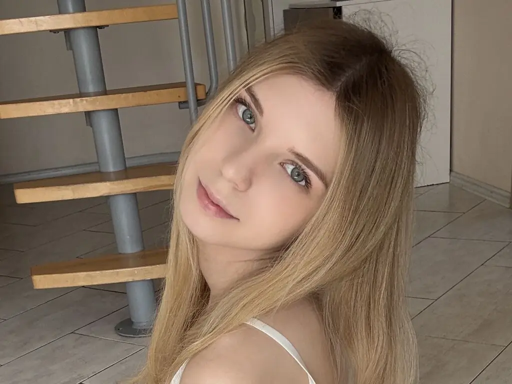 Blonde woman in a white tank top smiling in a modern home setting with wooden stairs.
