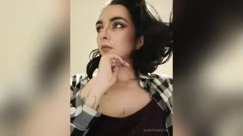 Moody portrait of a woman with dark hair wearing a plaid shirt and dark top in a minimalist indoor setting.