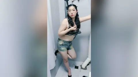 LouiseAnna poses in a white bathroom wearing a black lace bra and denim shorts, showing off her legs and midriff.