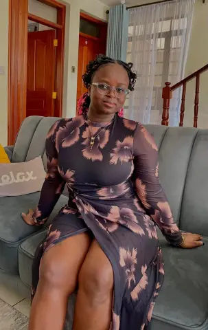 PrettyDoll22 wears a long-sleeved, dark floral print dress while sitting on a grey sofa, showing off her legs through a high slit.