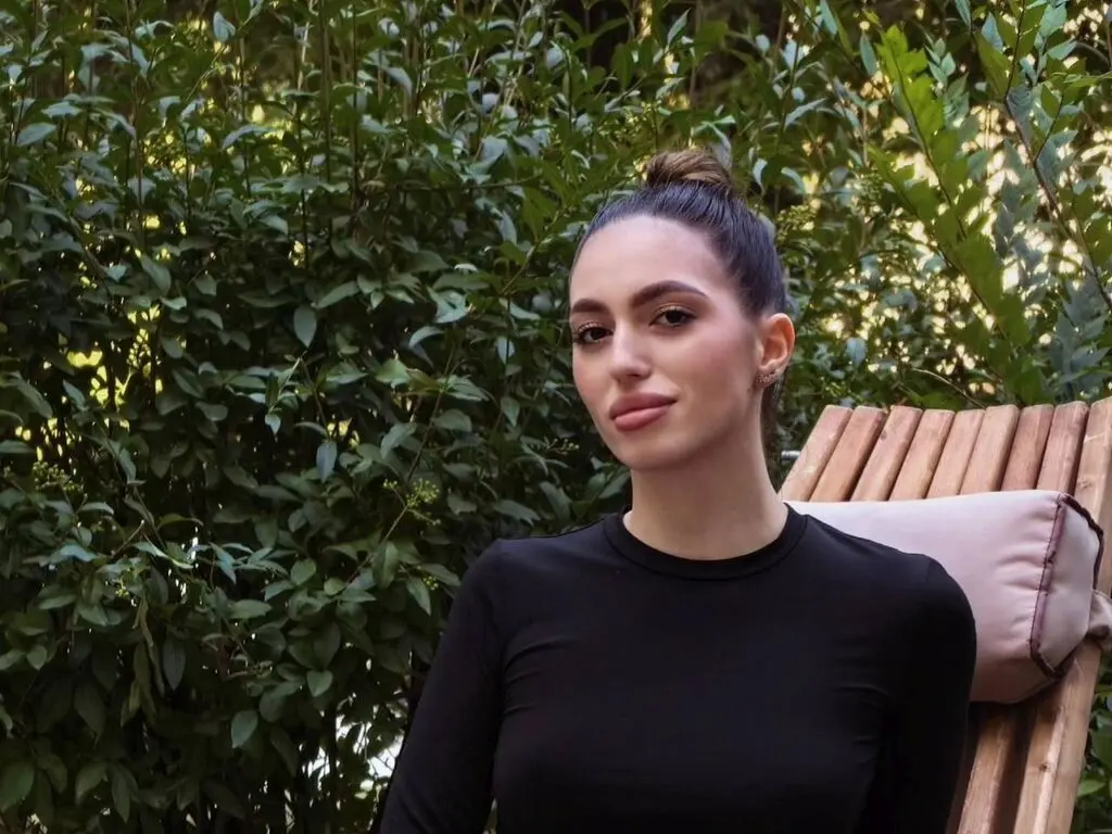 Young woman in a black long-sleeve top sitting on a wooden lounge chair in a lush green garden.