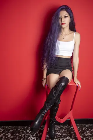Young woman with long purple hair in a white crop top and black shorts sitting against a red wall.