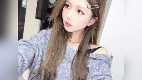 Young woman with long brown hair wearing a cozy grey sweater in a bright indoor setting.