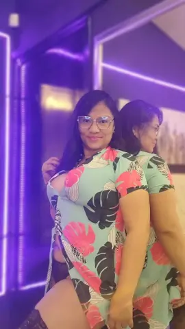 Smiling woman in a mint green floral print dress posing in a modern room with purple neon lighting.