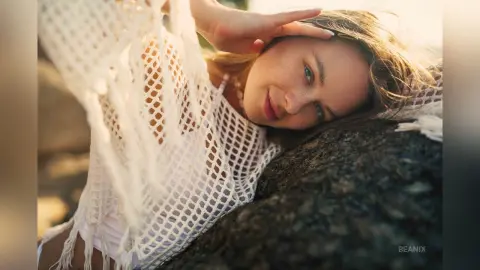 Smiling woman in a white crochet lace top relaxing on dark rocks during a warm sunset.