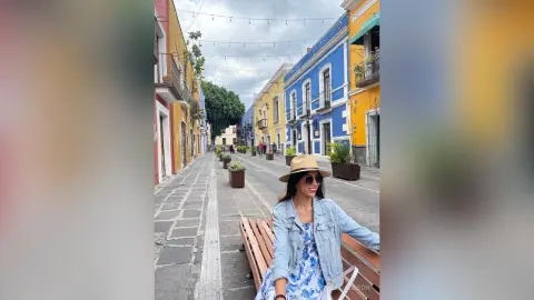 Smiling woman in a denim jacket and sun hat sitting on a bench in a colorful cobblestone street in Mexico.