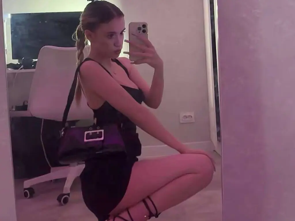 Young woman in a black slip dress and shoulder bag posing for a mirror selfie in a modern room.