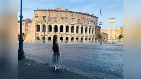 LucyMartin stands on a cobblestone street in front of a large, ancient stone amphitheater wearing a long, black and white striped dress.