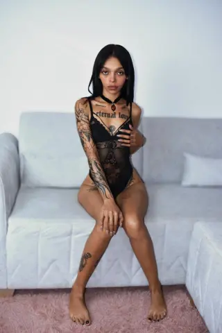emilyburton18 poses in a black lace bodysuit, showing off her tattoos and long dark hair while sitting on a light grey sofa.