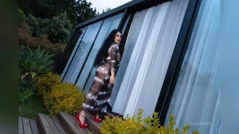 JuliahLewis stands outdoors in a long, sheer black and white patterned dress, posing next to large glass windows and green plants.