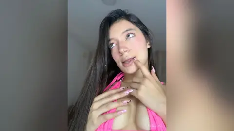 AleDilucas has long dark hair and pink blush, wearing a pink bikini top while touching her lips and showing cleavage.