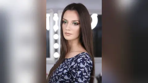 Young woman with long brown hair and green eyeshadow wearing a navy floral blouse in a bright studio setting.
