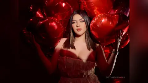 BrithneyDimons poses with large red heart-shaped balloons, wearing a red ruffled bustier top and showing her cleavage.