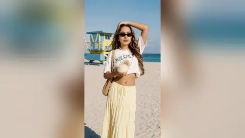 LilianMendoza poses on a sandy beach wearing a white t-shirt and long yellow skirt, holding a coconut while wearing sunglasses.