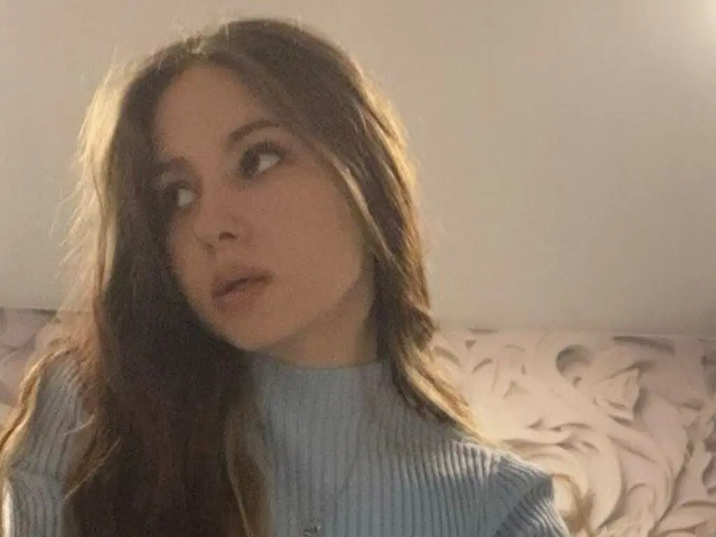 Young woman with long wavy hair wearing a blue ribbed turtleneck sweater in a cozy indoor setting.