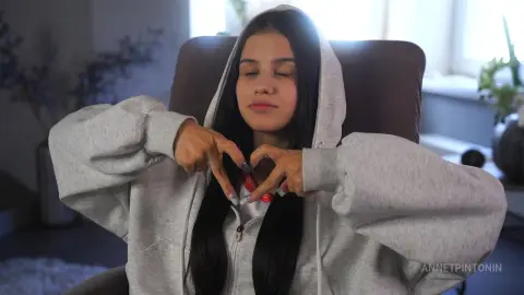 Young woman in a grey oversized hoodie making a heart gesture with her hands in a cozy indoor setting.