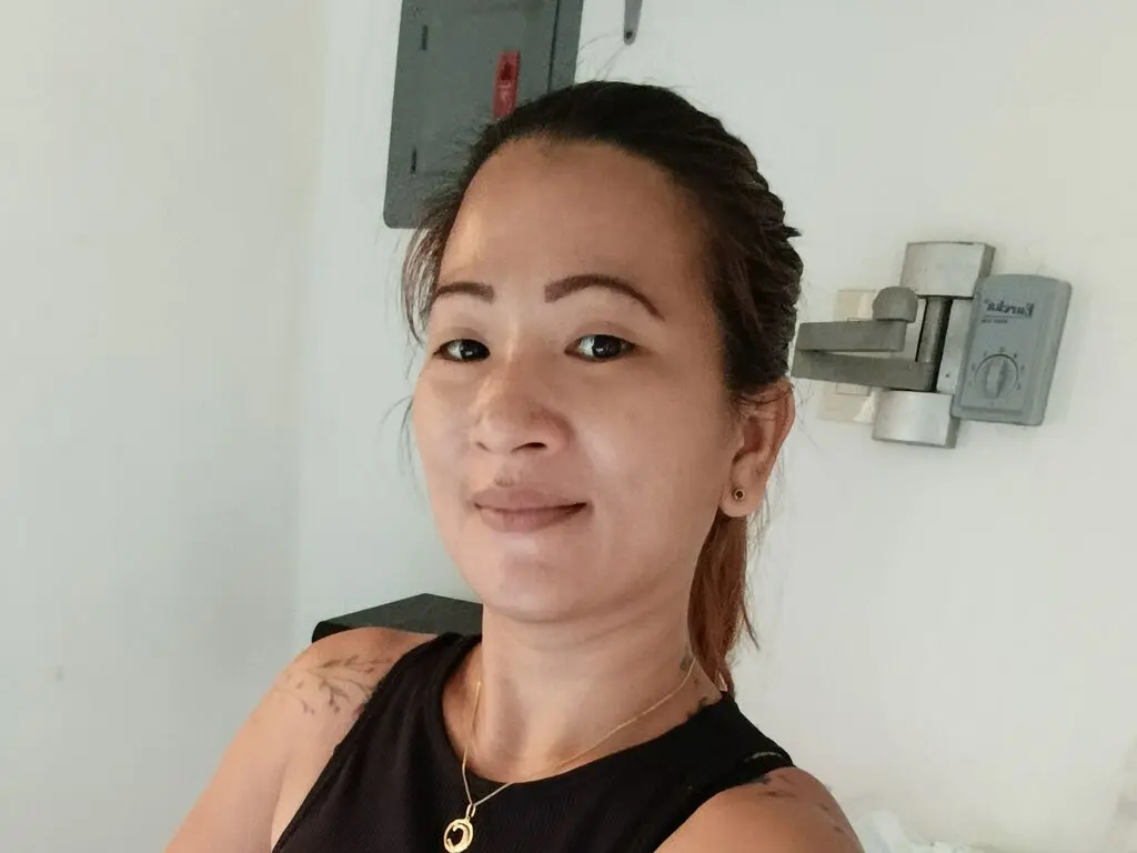 CristinaMimiss smiles at the camera wearing a black tank top and a gold necklace, showing her dark hair pulled back and a tattoo on her shoulder.