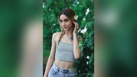 Young woman in a striped crop top and denim shorts posing in a lush green garden with white flowers.